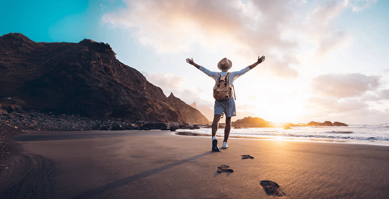 Man with a backpack and sun hat walking on a beach at sunrise, arms raised toward the sky