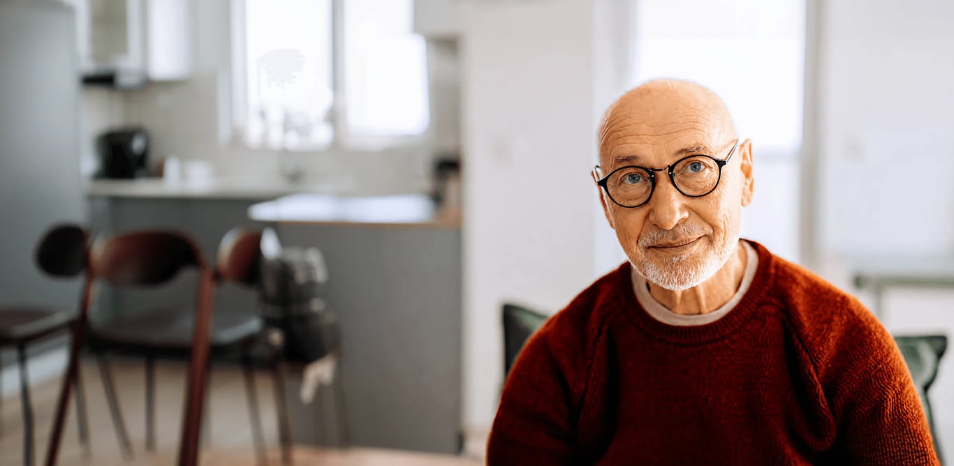 full-bleed photo of an older bald man with round black glasses and a white beard, wearing a rust-red sweater, smiling softly in a bright kitchen