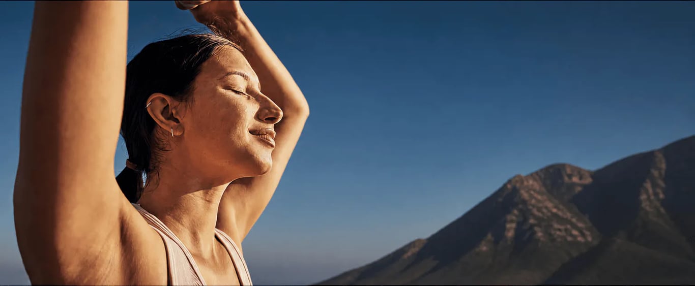 woman outdoors in sunlight against a blue sky, arm raised behind her head, eyes closed