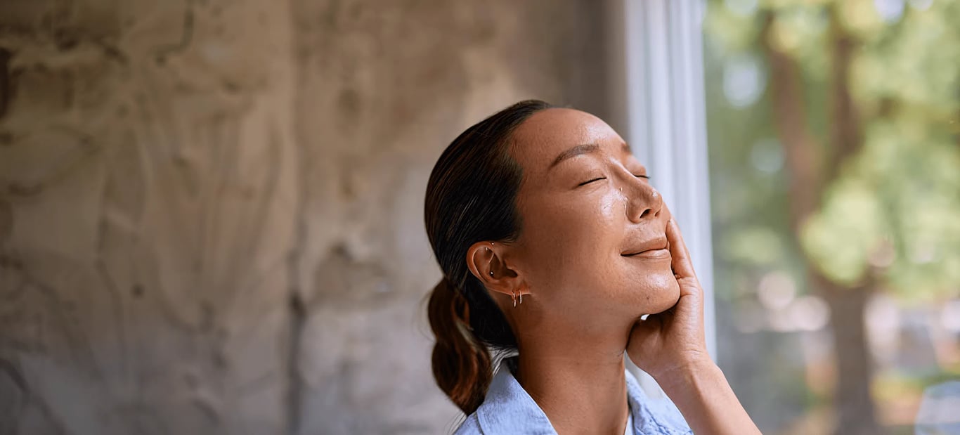 full-bleed photo of a woman with closed eyes touching her cheek, standing by a bright window with greenery outside