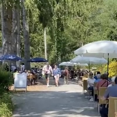 Lively garden cafe on sunny day. Visitors strolling on gravel path between seating areas and lush park vegetation.