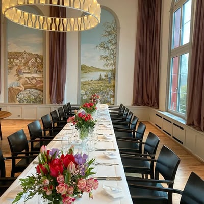 Gedecker Tisch mit farbenfrohen Tulpen- und Rosengestecken vor Wandgemälden in historischem Stil. Ringförmige Deckenleuchte und hohe Bogenfenster prägen den Raum.