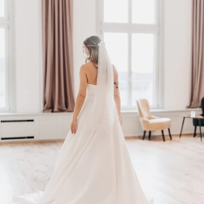 Braut in schlichtem weißen Hochzeitskleid steht im lichtdurchfluteten Festsaal mit hohen Fenstern und Rosé-Vorhängen.