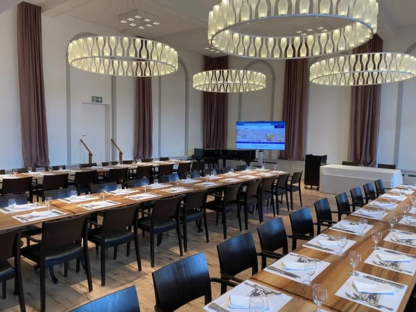 Conference setup with long wooden tables. Presentation screen and characteristic ceiling lights in elegant hall.