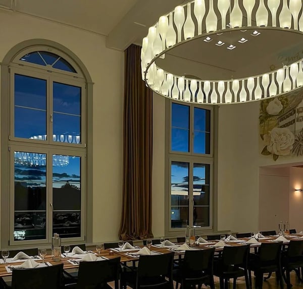 vening ambiance in the banquet hall featuring tall arched windows with a sunset view, elegant circular lighting fixture, and formally set dining tables.
