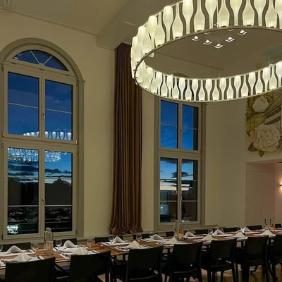 Abendstimmung im Festsaal mit hohen Bogenfenstern und Sonnenuntergangsblick, eleganter ringförmiger Beleuchtung und formal gedeckten Esstischen.