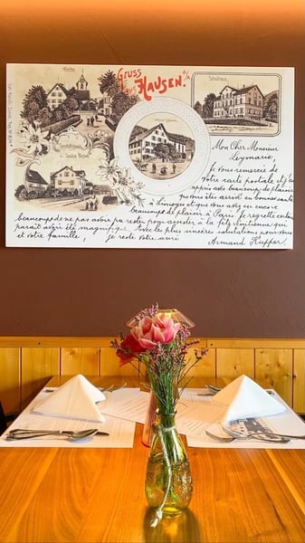 Traditional 'Stübli' lunch setting featuring a historic 'Gruss aus Hausen' postcard with multiple village scenes on the brown wall above. 