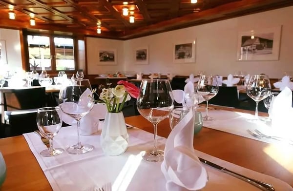 Bright dining room with wooden tables set with white linens, wine glasses and fresh flowers, historic photos on the walls, and a warm wooden ceiling.