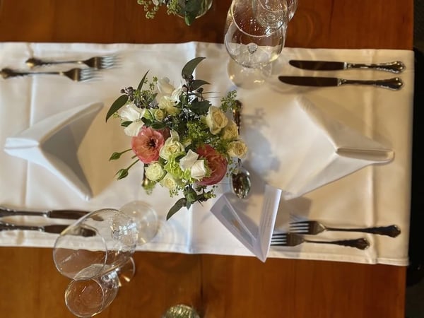 Formal table setting with a crisp white tablecloth and elegant place setting from above. The centerpiece features a delicate floral arrangement with pink ranunculus, cream roses, and fresh greenery in a glass vase.