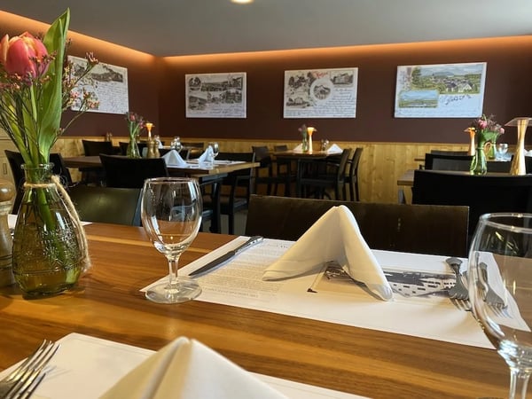 Restaurant table setting at eye level showing wooden tables with white napkins and wine glasses, historic postcards on the brown walls above wooden paneling, and fresh pink flowers in glass vases.
