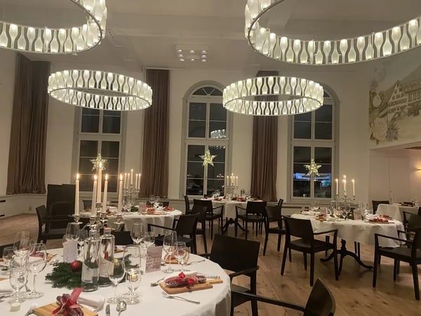 Löwen Hall at night with impressive ring-shaped lamps and Christmas decorations. Round tables formally set, candelabras and illuminated stars at arched windows.