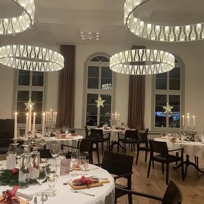 Löwen Hall at night with impressive ring-shaped lamps and Christmas decorations. Round tables formally set, candelabras and illuminated stars at arched windows.