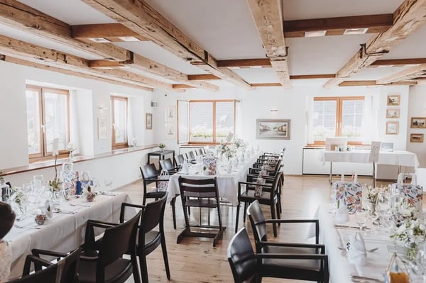 Formal dining room with wooden beam ceiling. White tables, black chairs and light floral decorations in bright space with wooden windows.