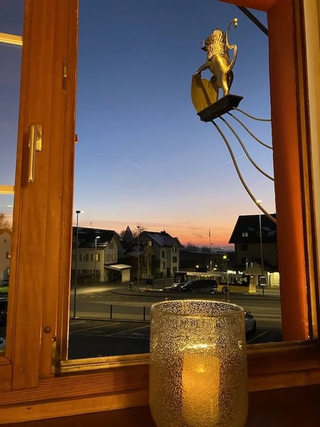 View from Restaurant Löwen window onto the Postplatz and bus station at dusk, with a golden lion emblem and candlelight in the foreground and a warm sunset sky over village buildings.