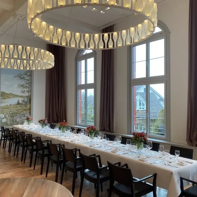 Lange Festtafel mit weissem Tischtuch und roten Blumengestecken. Landschaftsgemälde in historischem Stil und hohe Bogenfenster mit Ausblick.