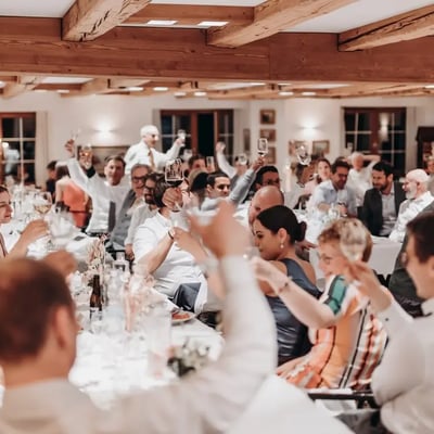 Festliche Hochzeitsgesellschaft hebt gemeinsam die Weingläser zum Toast in einem rustikalen Saal mit Holzbalkendecke.