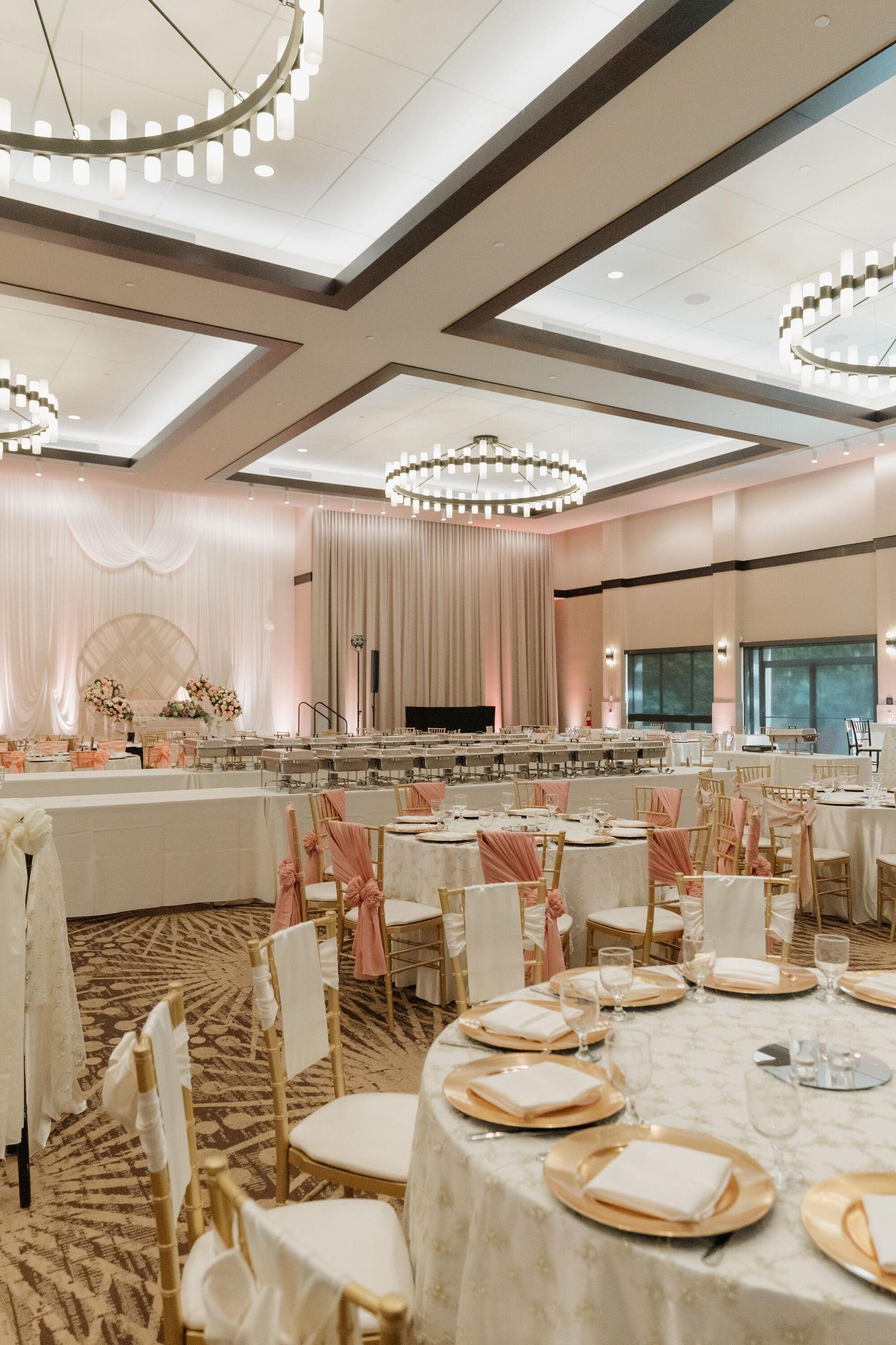 Grand Ballroom set with round tables, blush chair sashes, and place settings under ring chandeliers.