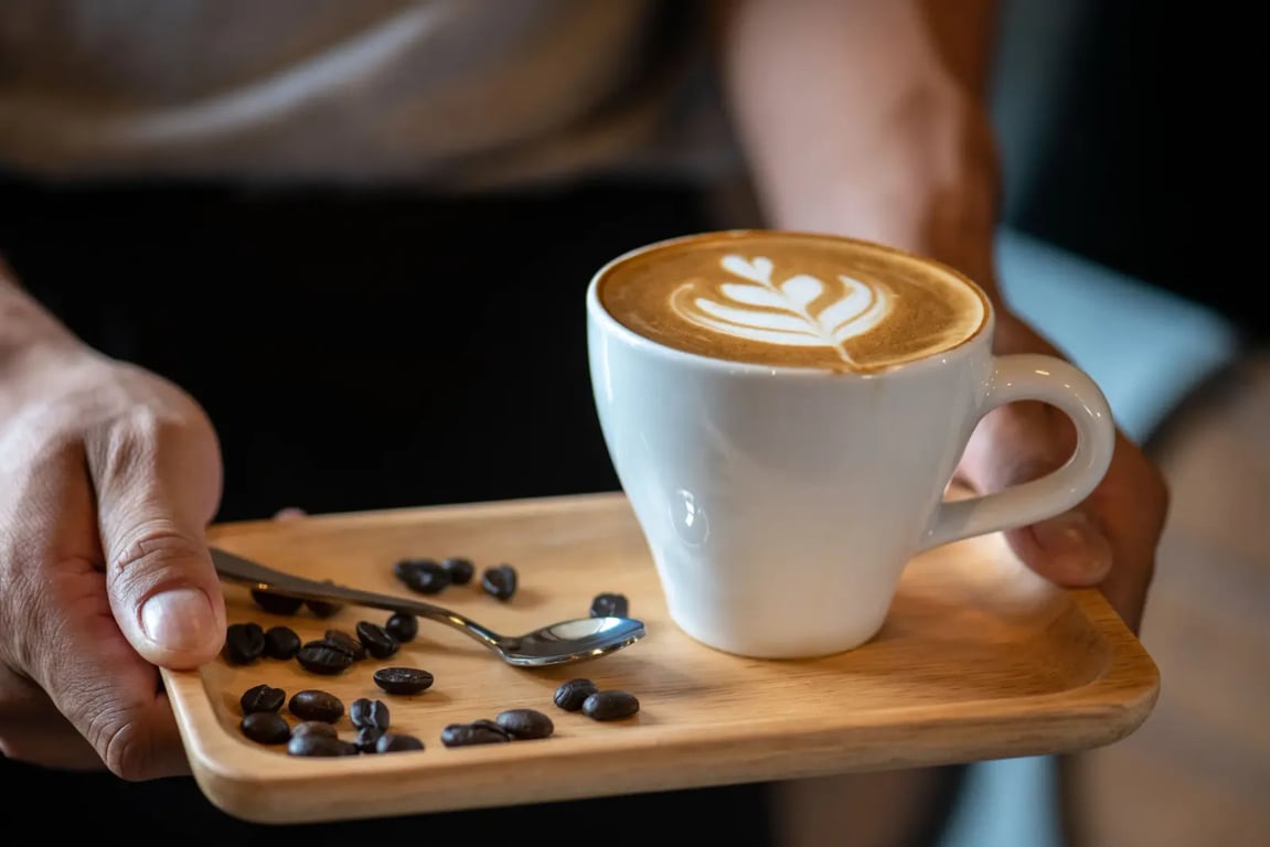 Žena v béžovém tričku a černé sukni drží menší dřevěný tácek. Na tácku jsou rozsypaná kávová zrna, je zde položená lžička a také bílá hrnek s kávou.