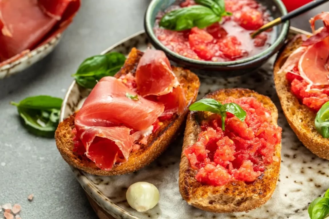 Detail na kousky opečeného chleba s nastrouhaným rajčetem, česnekem a plátky šunky na talíři s čerstvou bazalkou