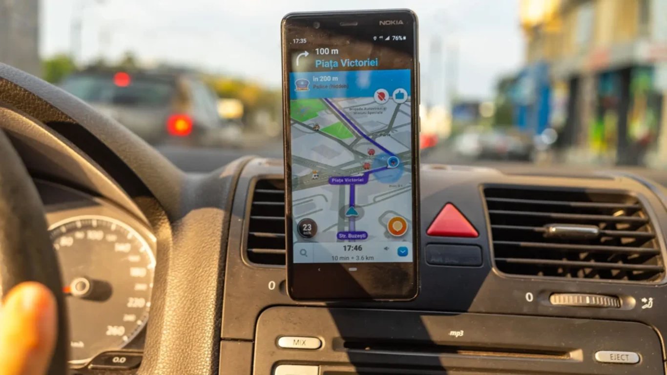 Mobilní telefon připevněný na palubní desce auta, na displeji běží navigace Waze s aktivní trasou a upozorněním na policii před řidičem.