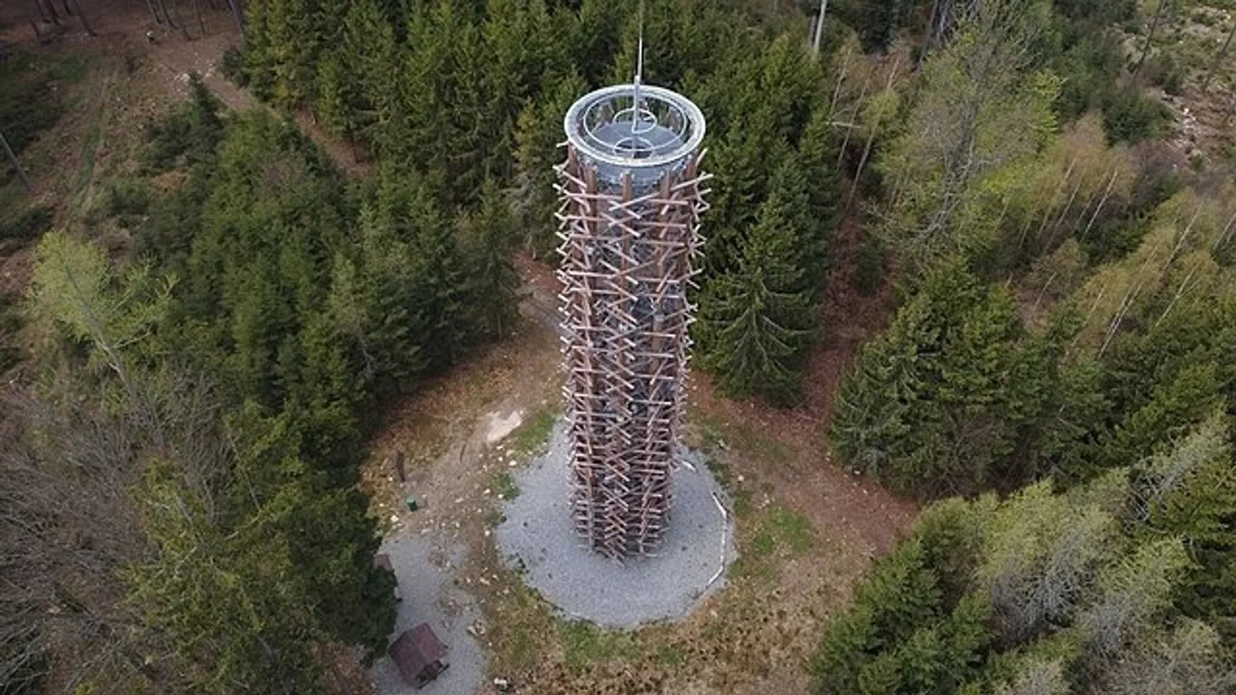 Uprostřed zelenajících se lesů na kulatém plácku, stojí rozhledna obložená dřevěnými trámy s kovovou konstrukci 
