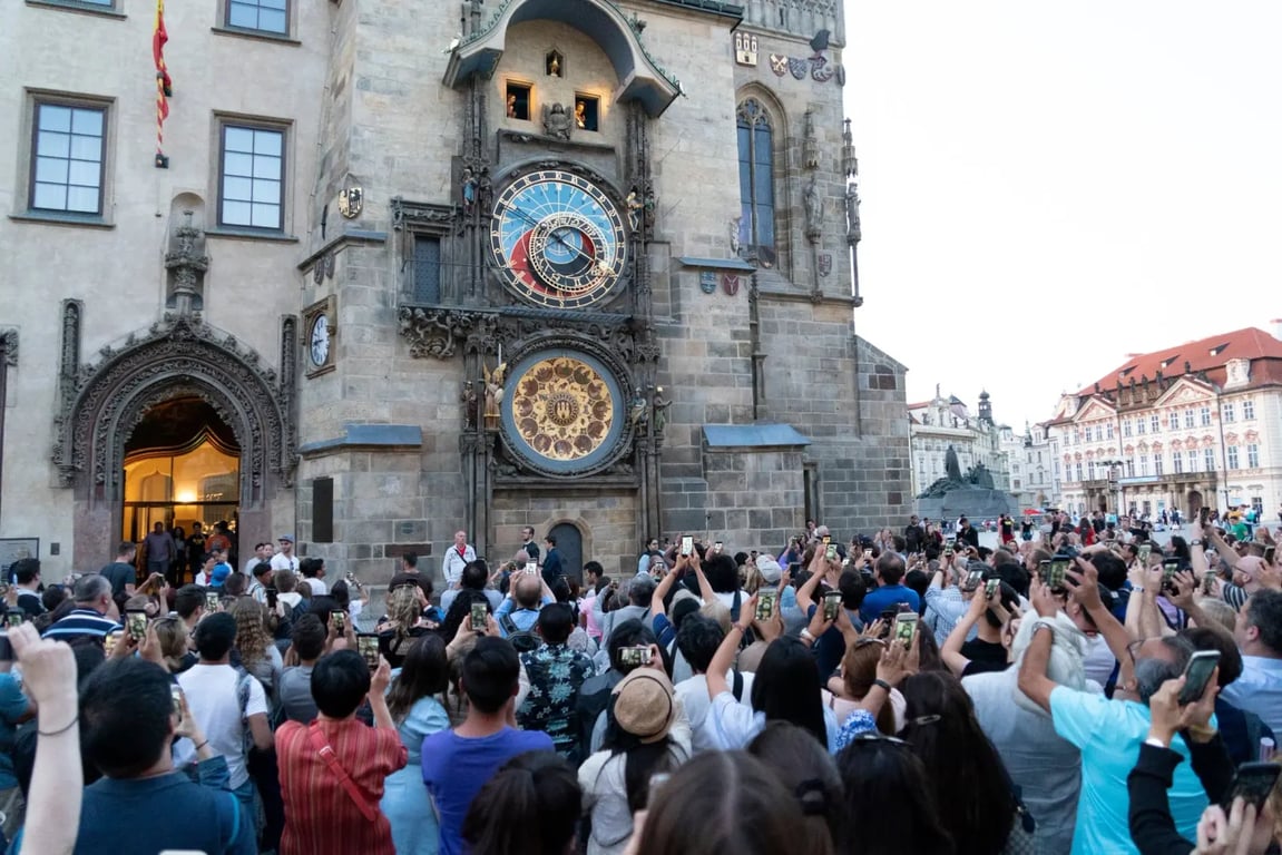 Dav turistů stojící před pražským orlojem. Je celá hodina, takže se objevují apoštolové, což si někteří turisté točí na své mobilní telefony.