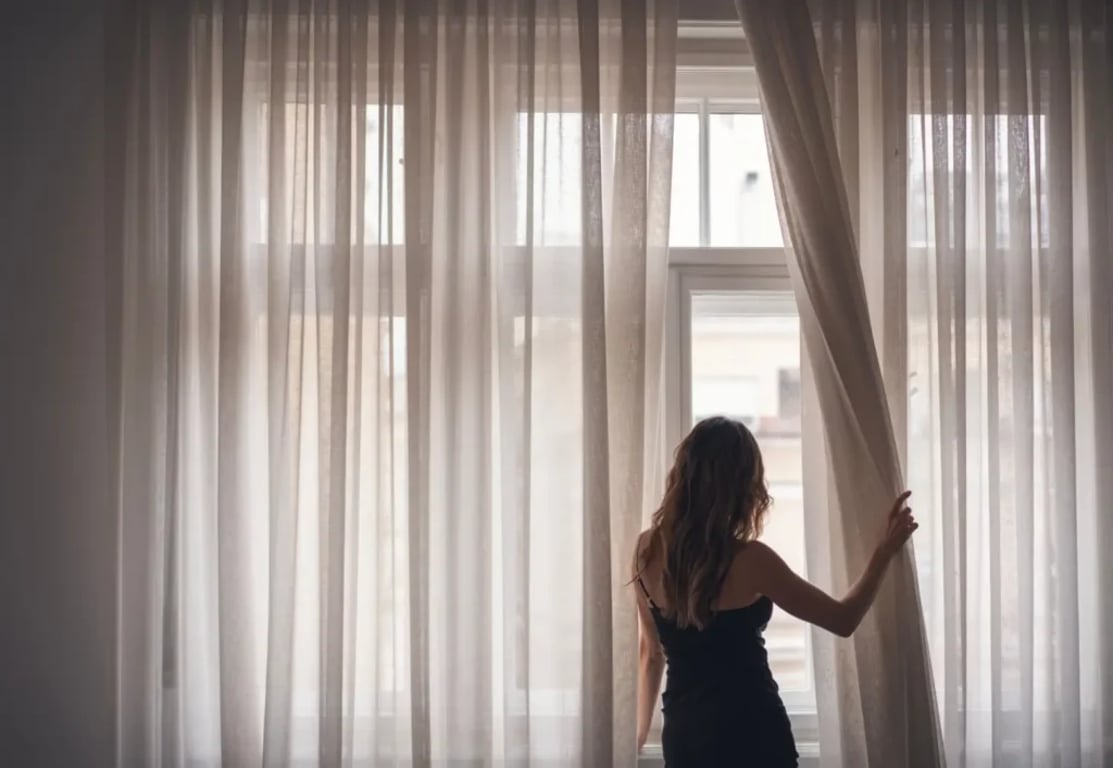 Žena ve tmavých šatech odhrnuje dlouhou průhlednou záclonu
