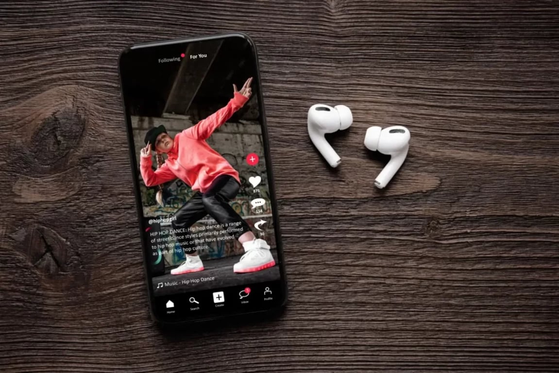 Smartphone na stole zobrazuje mladého tanečníka v červené mikině při natáčení videa na TikTok, vedle leží bezdrátová sluchátka