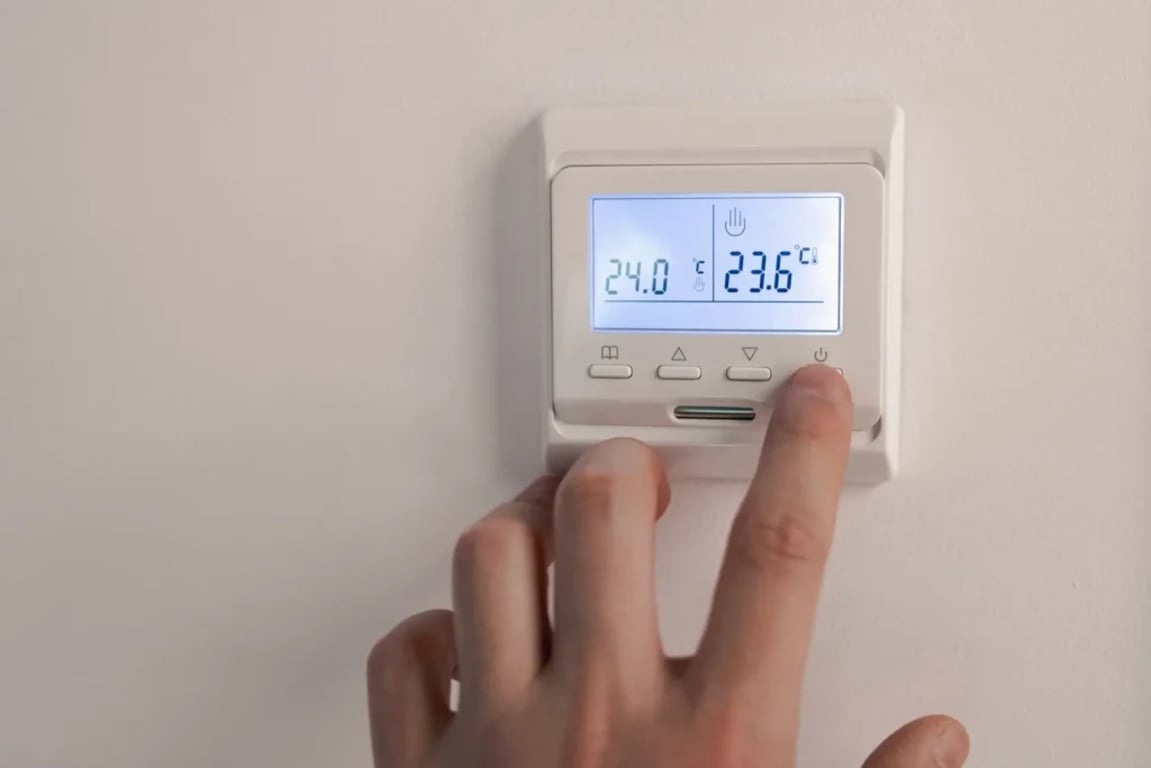Detail ruky, která mačká tlačítko na digitálním termostatu umístěném na zdi, displej zobrazuje aktuální a nastavenou teplotu v místnosti.