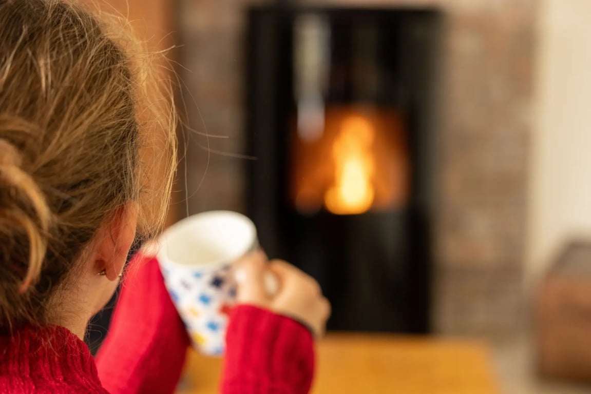Žena v červeném svetru sedí u kamen s hrnkem