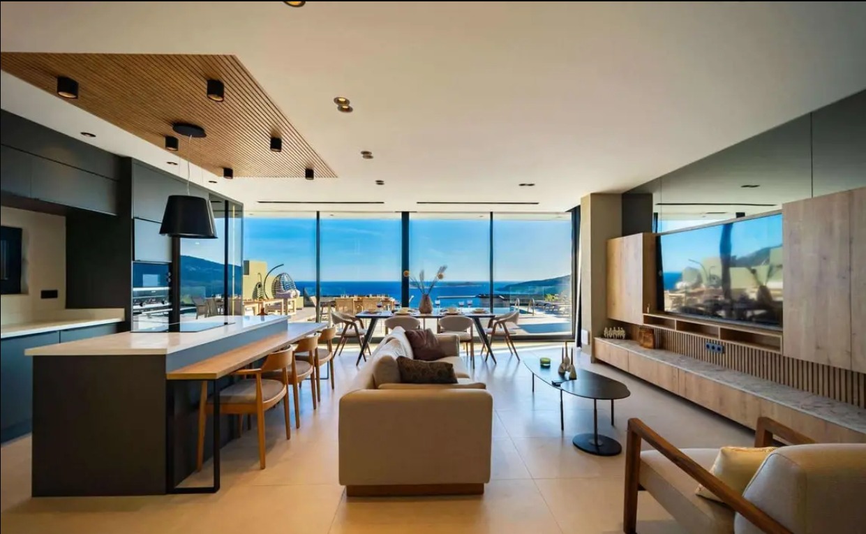 Modern ve geniş oturma alanına sahip, deniz manzaralı lüks villa iç mekanı. Açık mutfak, yemek masası, salon alanı ve geniş camlardan görünen Akdeniz ile Kalkan manzarası yer alıyor.