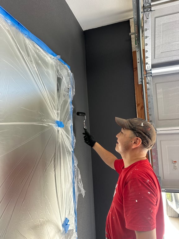Painter rolling dark gray paint on a garage wall, with plastic sheeting and blue painter's tape masking the area.