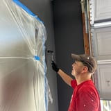 Painter rolling dark gray paint on a garage wall, with plastic sheeting and blue painter's tape masking the area.
