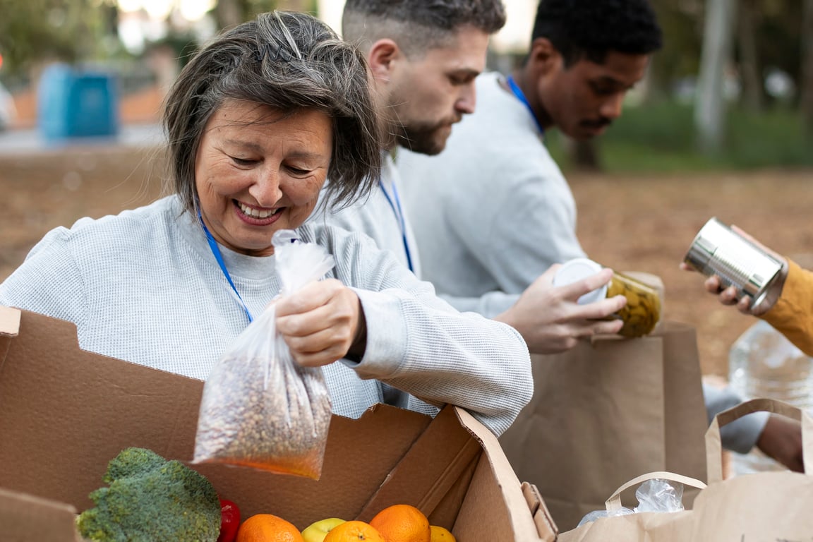 Smiling volunteer holding a bag of dried beans while sorting fresh produce and canned goods into boxes during a community food distribution event outdoors.