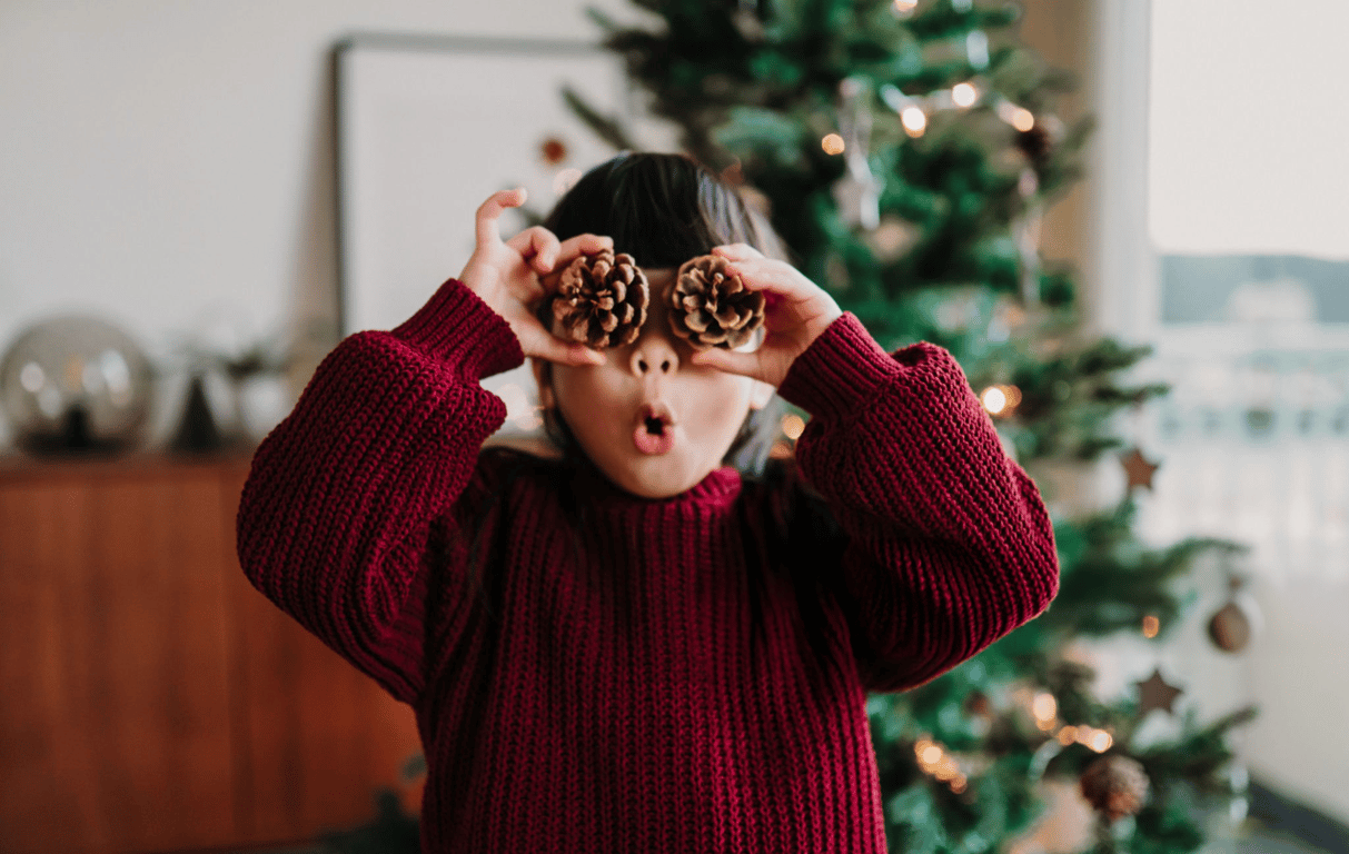 Child in a cozy red sweater holding pinecones over their eyes, making a playful face in front of a decorated Christmas tree.