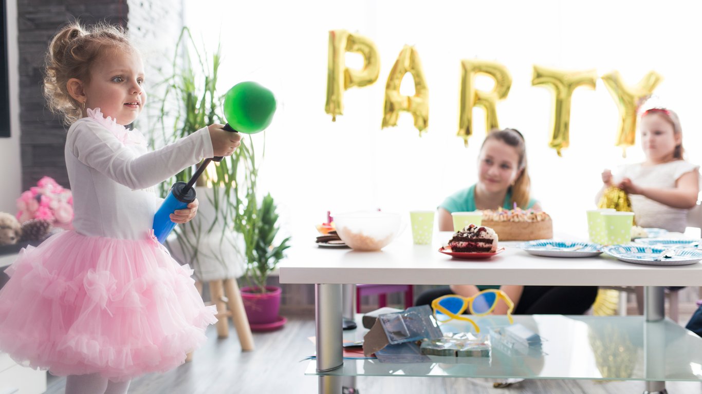 Young girl in a pink tutu playing at a kids’ birthday party with friends, balloons, cake, and gold party decorations in the background.