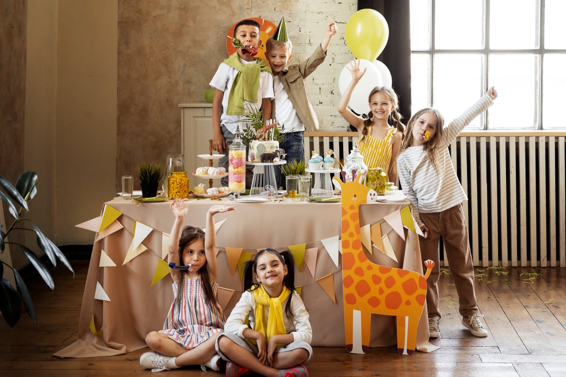 There is a table with cakes and sweets with children facing towards the camera celebrating.