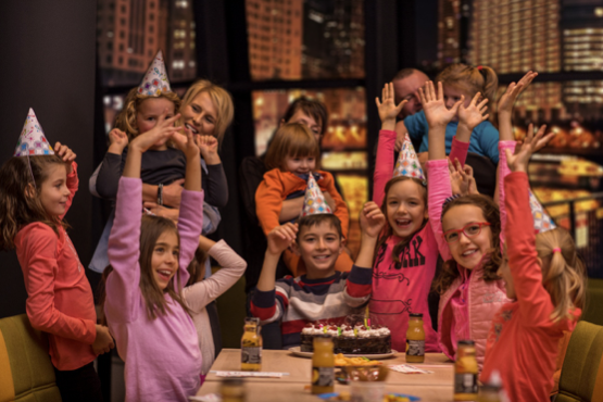 A group of young children celebrating a birthday party sat around a table with their arms up. On the table there is a cake and juice and all children are wearing birthday party hats.