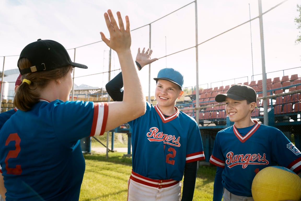 Children in blue baseball uniforms smiling and giving high fives on a baseball field.