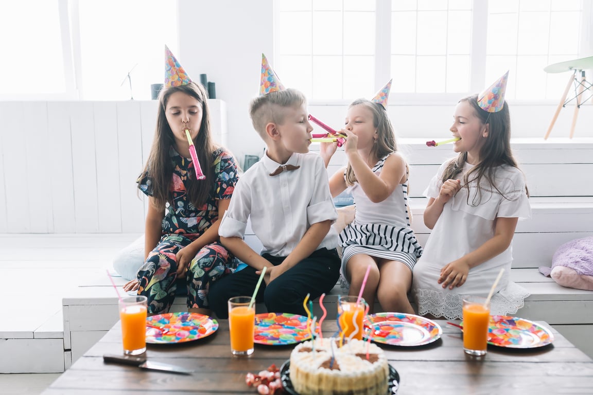 boys and girls sat on wooden bench with drinks and plates, blowing party horns near cake