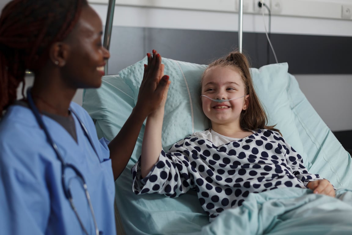Smiling nurse and young girl in a hospital bed share a high five. The child is wearing a nasal cannula and hospital gown, while smiling and happy.