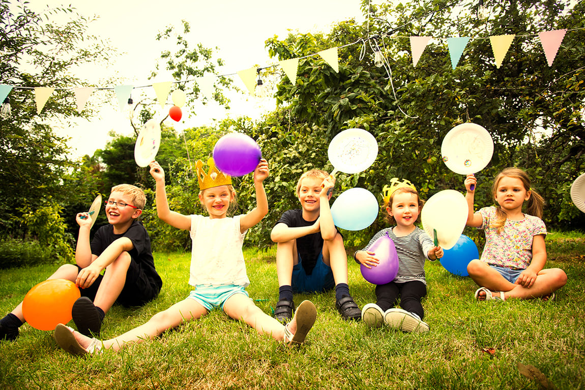 Children sitting on grass at a summer birthday party holding balloons and paper plates, with bunting and string lights hanging in the background.