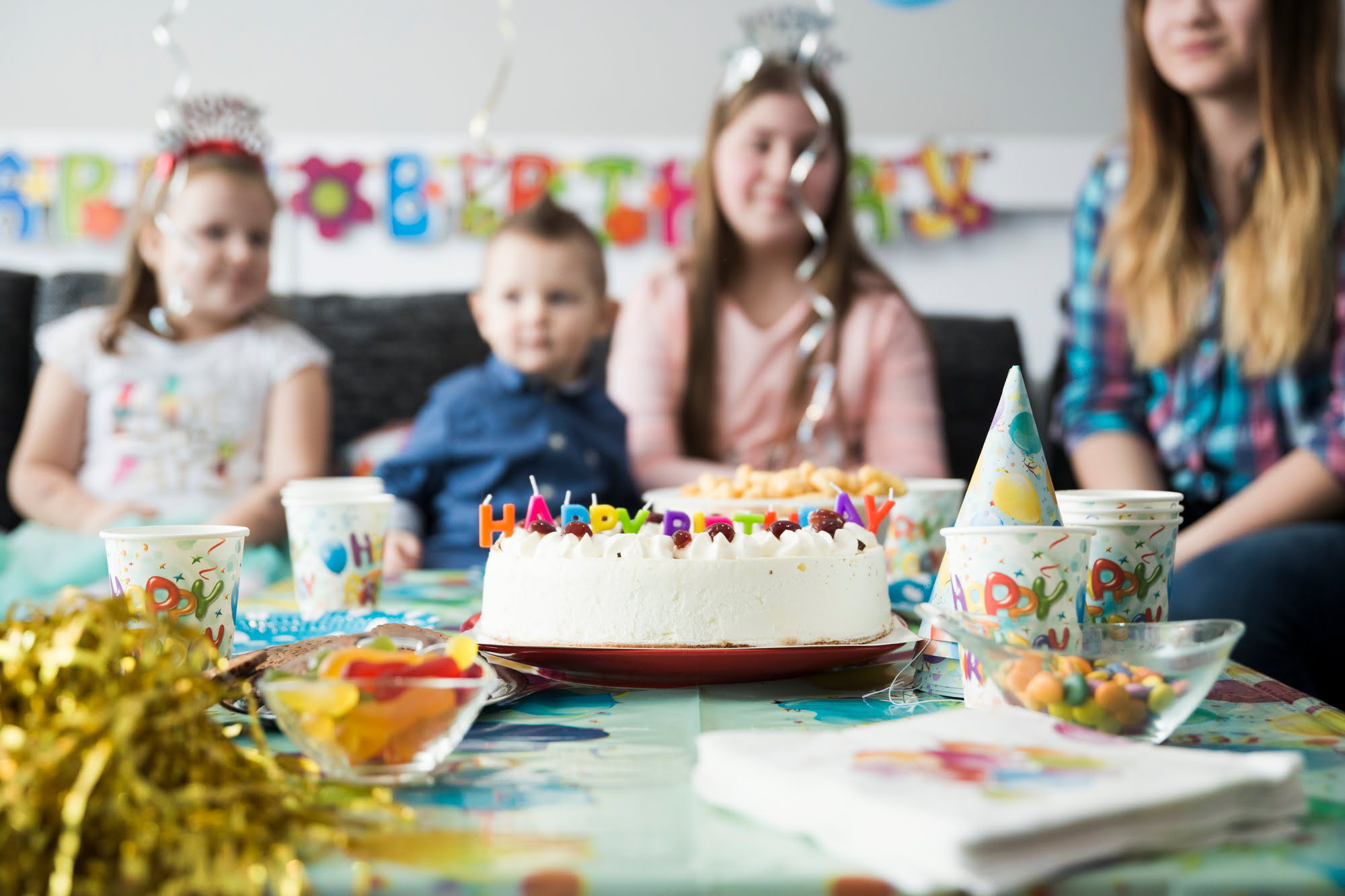 Children gathered around a birthday party table with a white cake decorated with Happy Birthday candles, party cups, and sweets in the foreground.