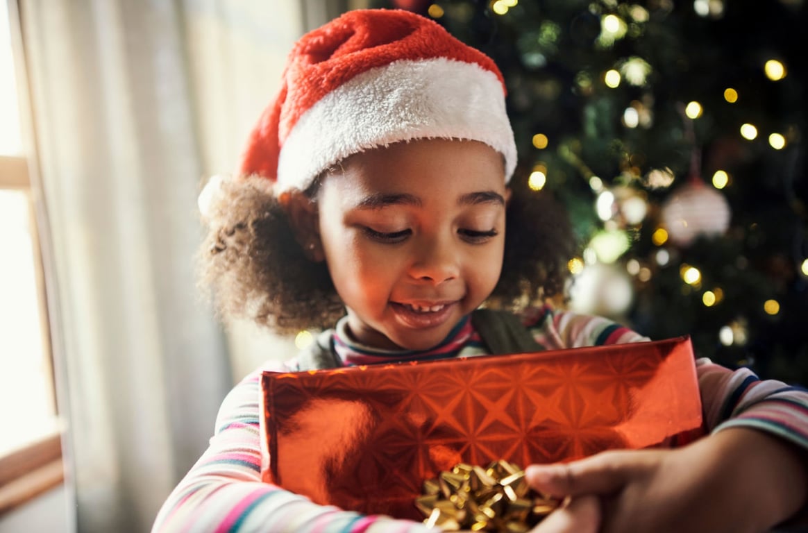 Child in a cozy red sweater holding pinecones over their eyes, making a playful face in front of a decorated Christmas tree.
