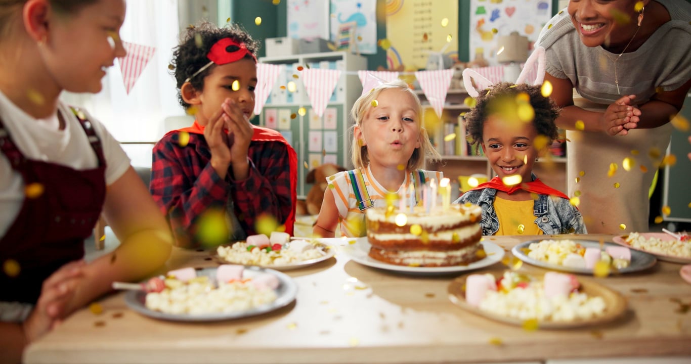 Children around a classroom table watching a child blow out her birthday cake candles.