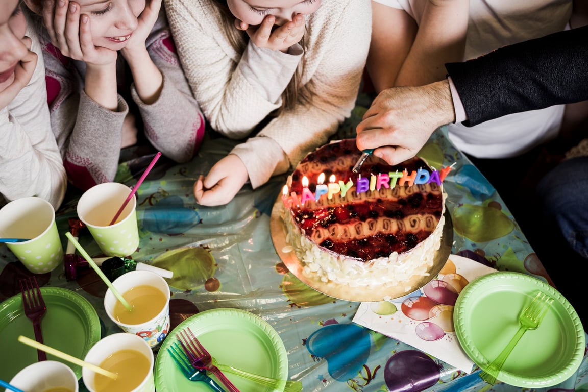 high angle shot of a birthday party celebration. There are a group of girls sat around a table watching someone light the candles on a cake that reads “happy birthday”