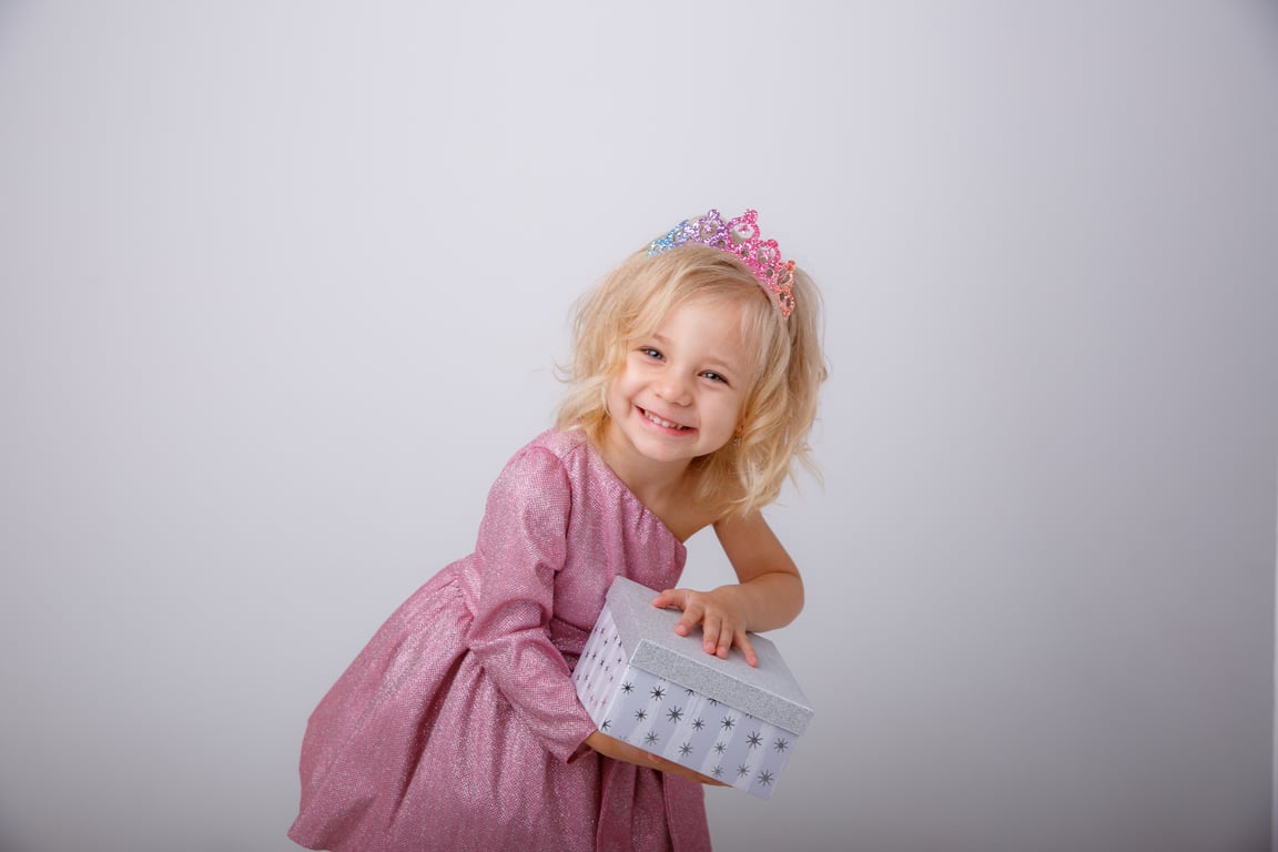 Smiling young girl in a sparkly pink princess dress and colourful tiara holding a gift box against a light grey background.