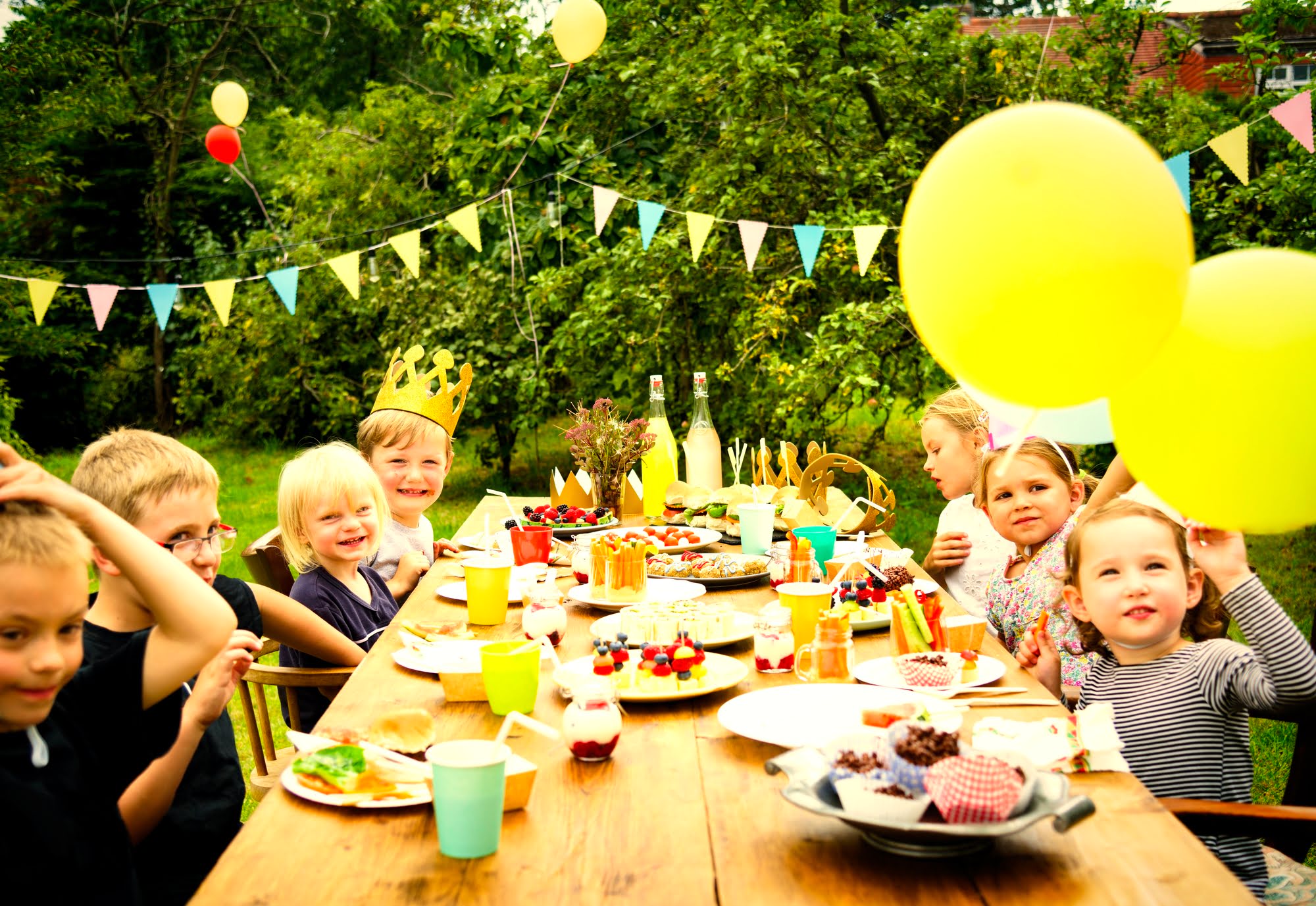 Children sitting around a wooden outdoor table at a backyard birthday party, with balloons, bunting, and a spread of food and drinks in a garden setting.