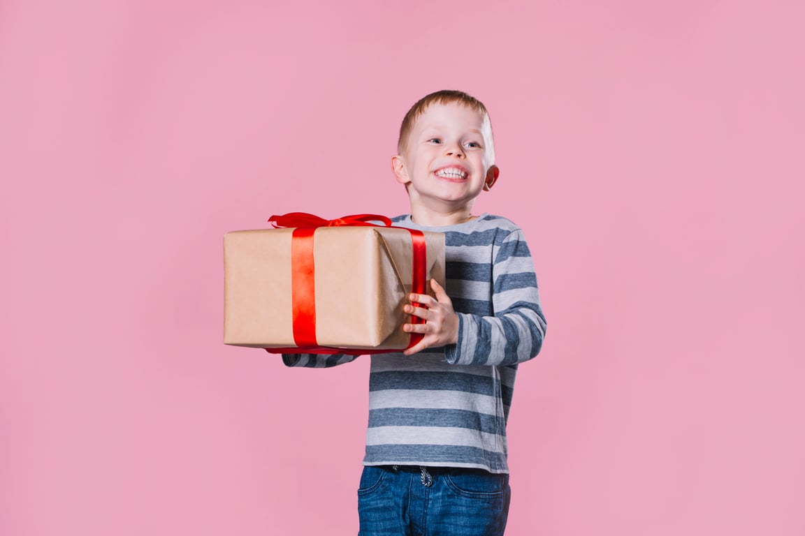 A child smiling while holding a large present against a pink background.