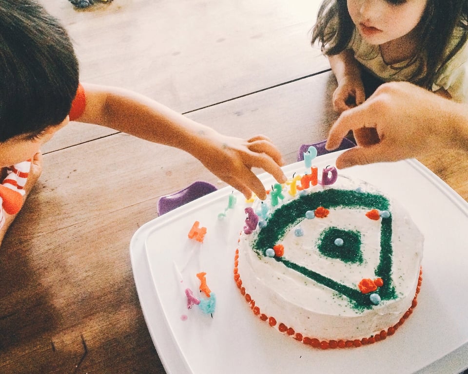 Children gathered around a birthday party table with a white cake decorated with Happy Birthday candles, party cups, and sweets in the foreground.